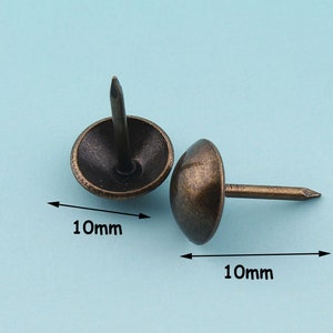 May include: Two antique gold-toned tacks with round heads, each measuring 10mm in diameter. The tacks have a pointed nail.