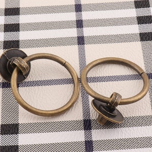 Puede incluir: Dos tiradores de puerta de armario de latón antiguo con un diseño de anillo redondo. Los tiradores tienen un pomo decorativo en la parte inferior.