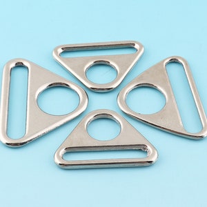Pode incluir: Quatro anéis metálicos triangulares prateados com uma abertura arredondada. Os anéis são projetados para serem fixados em tecido ou outros materiais.
