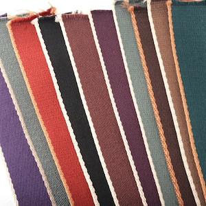 Puede incluir: Una colección de diez tiras de tela en varios colores, incluyendo morado, gris, naranja, rojo, negro, marrón y azul verdoso. Cada tira tiene una franja blanca que recorre el centro.