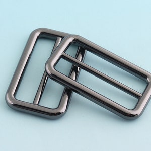 Puede incluir: Dos deslizadores de hebilla de cinturón de metal negro. Los deslizadores son rectangulares con esquinas redondeadas y tienen un acabado liso y brillante.