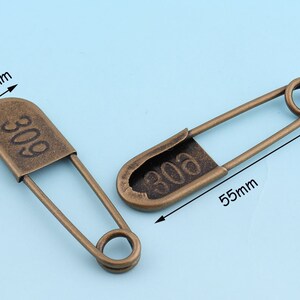 May include: Two antique gold safety pins with the number "306" engraved on them. The pins are 5.5 inches long and 1.3 inches wide.