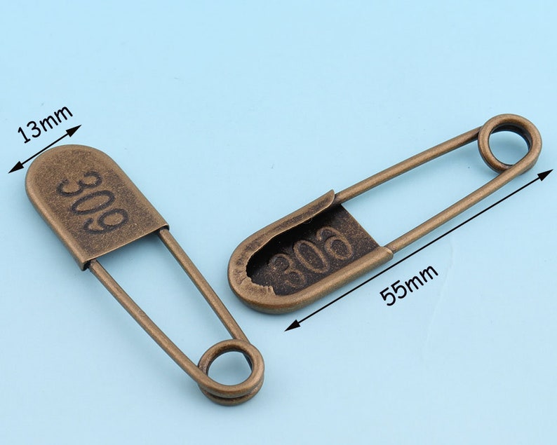 May include: Two antique gold safety pins with the number "306" engraved on them. The pins are 5.5 inches long and 1.3 inches wide.