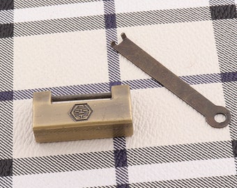 Puede incluir: Un candado de latón con un ojo de cerradura y un diseño decorativo en la parte delantera. El candado está sobre una superficie a cuadros blancos y negros. Una llave de latón está al lado del candado.