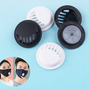 Könnte beinhalten: Fünf runde, weiße und schwarze Kunststoff-Ventilkappen für Gesichtsmasken. Die Kappen haben ein Gittermuster und sind so konzipiert, dass sie an der Außenseite einer Gesichtsmaske befestigt werden.