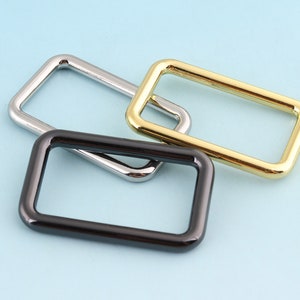 Op de afbeelding: Drie metalen riemgespen in verschillende afwerkingen: zilver, zwart en goud. De gespen zijn rechthoekig en hebben afgeronde hoeken.
