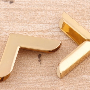 Op de afbeelding: Twee gouden metalen hoekbeschermers. De hoekbeschermers zijn L-vormig en hebben een gladde, glanzende afwerking.