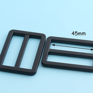 Puede incluir: Dos deslizadores de hebilla de plástico negro, uno de 45 mm de largo. Las hebillas tienen una forma rectangular con dos ranuras paralelas.