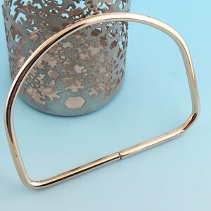 May include: A gold-colored, geometric bracelet with a D-shaped design. The bracelet is made of a shiny metal and has a clasp closure. The background is a light blue surface with a decorative metal candle holder.