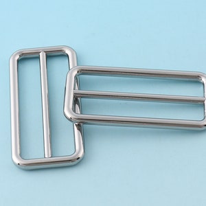 Puede incluir: Dos hebillas deslizantes de metal plateado. Las hebillas son rectangulares con esquinas redondeadas y tienen una sola ranura en el centro.
