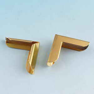 May include: Two gold-colored, L-shaped metal corner protectors. The protectors have a shiny finish and are designed to fit over the corners of a box or frame. They are set against a light blue background.