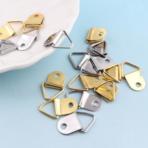 May include: A collection of gold and silver triangle-shaped picture hanging hooks. The hooks have a small loop at the top for hanging a picture or other object.
