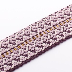 May include: Close-up of a woven fabric strip with a repeating floral pattern. The strip features a combination of purple, lavender, and cream colors, with thin gold threads running through the center. The edges are a darker purple.