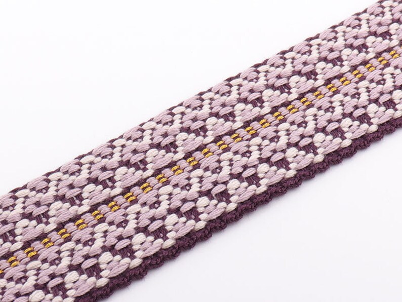 May include: Close-up of a woven fabric strip with a repeating floral pattern. The strip features a combination of purple, lavender, and cream colors, with thin gold threads running through the center. The edges are a darker purple.