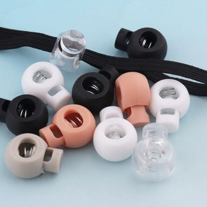 Może przedstawiać: Zestaw plastikowych zatrzasków do sznurka w różnych kolorach, w tym przezroczystym, białym, czarnym, brązowym i beżowym. Każdy zatrzask ma mechanizm sprężynowy i okrągły kształt.