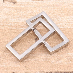 Puede incluir: Dos anillos rectangulares de metal plateado, uno ligeramente más grande que el otro, están apilados uno encima del otro. Los anillos son lisos y brillantes.