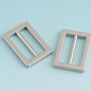 Puede incluir: Dos hebillas deslizantes de metal plateado. Las hebillas son rectangulares con un acabado liso y pulido.