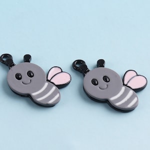 Puede incluir: Dos abalorios de abeja grises con ojos negros y alas rosas. Las abejas tienen un patrón de rayas blancas en sus cuerpos y un lazo negro para sujetar.