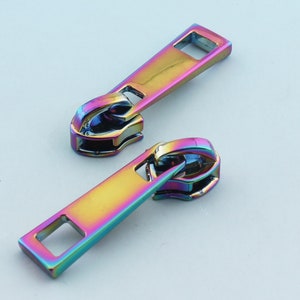 Peut inclure: Deux tirettes de fermeture éclair irisées avec un curseur rectangulaire et une boucle en métal. Les tirettes sont arc-en-ciel de couleurs, y compris le rose, le bleu, le vert et le jaune.