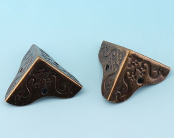 May include: Two antique brass corner protectors with an ornate floral design. The protectors are triangular in shape and have a distressed finish.
