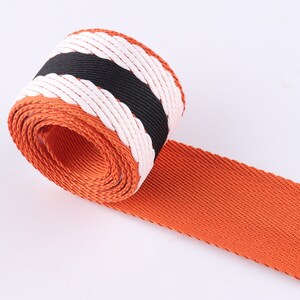 May include: Orange, white, and black striped webbing with a woven texture. The webbing is rolled up and the orange side is facing the camera.