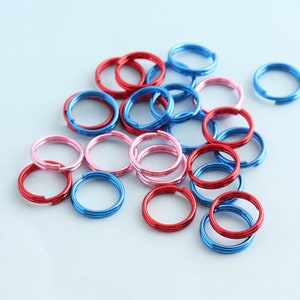 Könnte beinhalten: Sortiment aus bunten Metall-Sprühringen in Rot, Blau und Pink. Die Ringe sind auf einem hellblauen Hintergrund gestapelt und verstreut.