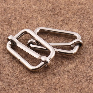 Puede incluir: Dos hebillas deslizantes de metal plateado para correas o cinturones. Las hebillas son rectangulares con esquinas redondeadas y tienen un acabado liso y pulido.