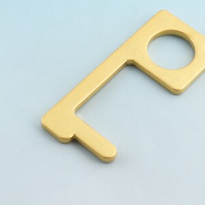 May include: A gold-colored, metal no-touch door opener and button pusher. The tool has a rectangular shape with a circular opening and a hook-like extension. The item is designed to minimize contact with surfaces.