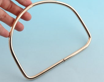 May include: A gold-colored, semi-circular handbag handle. The handle is made of a smooth, metallic material and has a rectangular base. The handle is held by a hand against a light blue background.