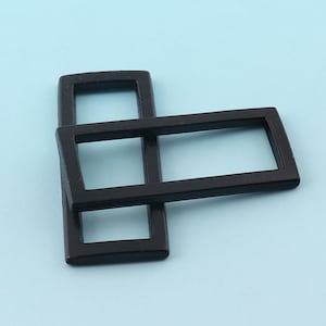 May include: Two interlocking, rectangular, black metal buckles against a light blue background. The buckles are made of a solid, matte material. The design features a smaller square buckle interlocked with a larger rectangular buckle.