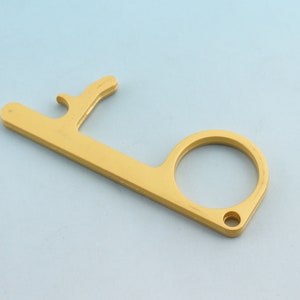 May include: A gold-colored touch tool with a finger loop and a hook-shaped end. The tool is designed to help users avoid direct contact with surfaces. The tool is made of metal and has a smooth finish.