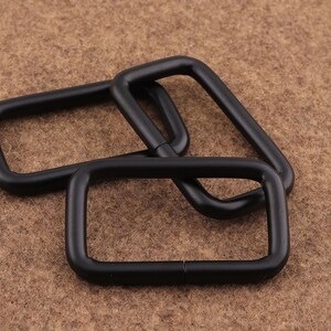 May include: Three black metal D-rings, also known as D rings or D-shaped rings, are stacked on top of each other. The rings are smooth and have a matte finish.