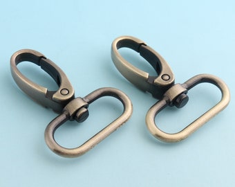 May include: Two antique gold-toned metal swivel clasps with a D-ring. The clasps are closed and ready to be attached to a strap or chain.