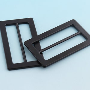 Puede incluir: Dos hebillas de cinturón rectangulares negras con una barra central. Fabricadas en material negro mate, destacan sobre un fondo azul claro. Estas hebillas están diseñadas para cinturones o correas.
