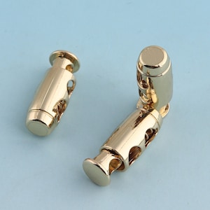 May include: Two gold-toned metal cord stoppers. The stoppers have a cylindrical shape with a series of holes around the middle. The stoppers are designed to secure cords or straps.