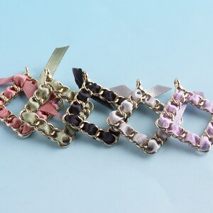 Op de afbeelding: Vier gouden ketting vierkante frames met verschillende gekleurde lint accenten. De frames zijn roze, groen, zwart en paars.