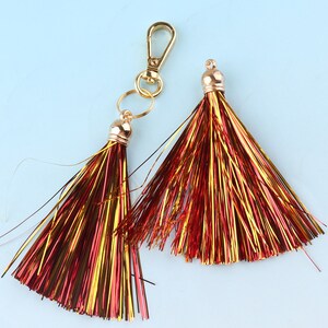 Può includere: Due portachiavi con nappe dorate con fili metallici rossi e dorati. Le nappe sono attaccate ad anelli dorati e una chiusura dorata.