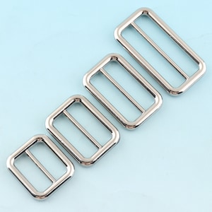 Puede incluir: Cuatro hebillas deslizantes de metal plateado de diferentes tamaños. Las hebillas son rectangulares y tienen un acabado liso.