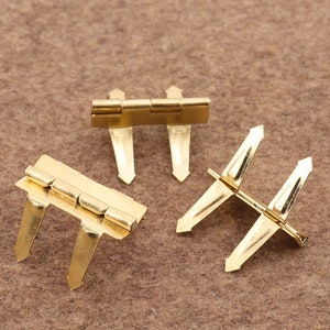 Puede incluir: Tres bisagras de metal dorado, cada una con tres puntas. Las bisagras están diseñadas para unir dos piezas de material.