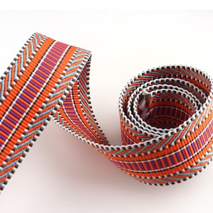 May include: Orange, purple, and blue patterned ribbon with a chevron design. The ribbon is rolled up and ready to be used for crafting or decorating.