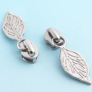 Könnte beinhalten: Zwei silberne Reißverschluss-Zipper mit einem Blattdesign. Die Zipper sind aus Metall und haben eine glänzende Oberfläche.