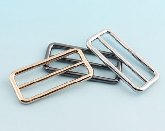 May include: Three metal slider buckles in gold, silver, and black. The buckles are rectangular with two parallel bars across the center.