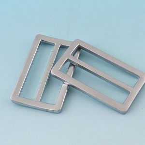Puede incluir: Dos hebillas rectangulares plateadas con un diseño de tres barras. Las hebillas son de metal mate y están sobre un fondo azul claro. Probablemente se utilizan para cinturones o correas.