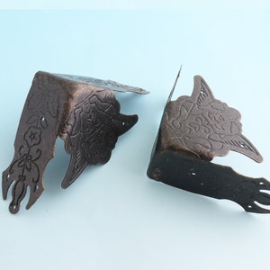 Peut inclure: Deux équerres d'angle antiques de couleur bronze avec des motifs floraux et de volutes. Chaque équerre a une forme en angle droit avec des découpes décoratives et des trous de montage. Ces équerres sont probablement pour des meubles ou des projets décoratifs.