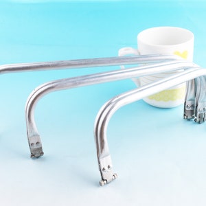 Könnte beinhalten: Mehrere silberfarbene Metallrahmen für Handtaschen, gebogen und gewölbt, mit Verschlussmechanismen, sind auf einer hellblauen Oberfläche angeordnet. Im Hintergrund befindet sich eine weiße Tasse mit gelben Blumenmustern.