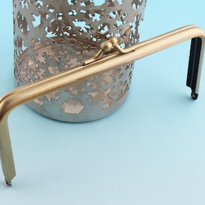 Puede incluir: Un marco de bolso de metal dorado con una correa negra. El marco está diseñado para ser fijado a un bolso o una bolsa.