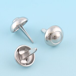 Puede incluir: Tres tachuelas de metal plateado en forma de cúpula con un extremo puntiagudo. Las tachuelas están sobre un fondo azul claro.