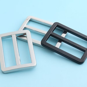 Puede incluir: Tres hebillas correderas rectangulares de metal en plata y negro. Las hebillas tienen un marco rectangular con dos barras paralelas en el interior. El fondo es azul claro.
