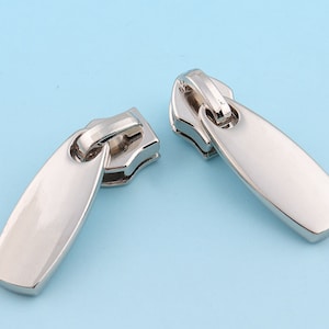 May include: Two polished silver zipper pulls, each with a rounded rectangular form and a small loop at the top. They are displayed against a light blue backdrop, accentuating their metallic lustre and design.