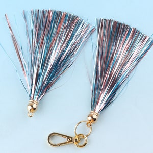 May include: Two decorative tassels with metallic strands in shades of blue, silver, and copper. Each tassel has a gold-colored cap and one has a gold-colored clasp. The tassels are displayed on a light blue background.